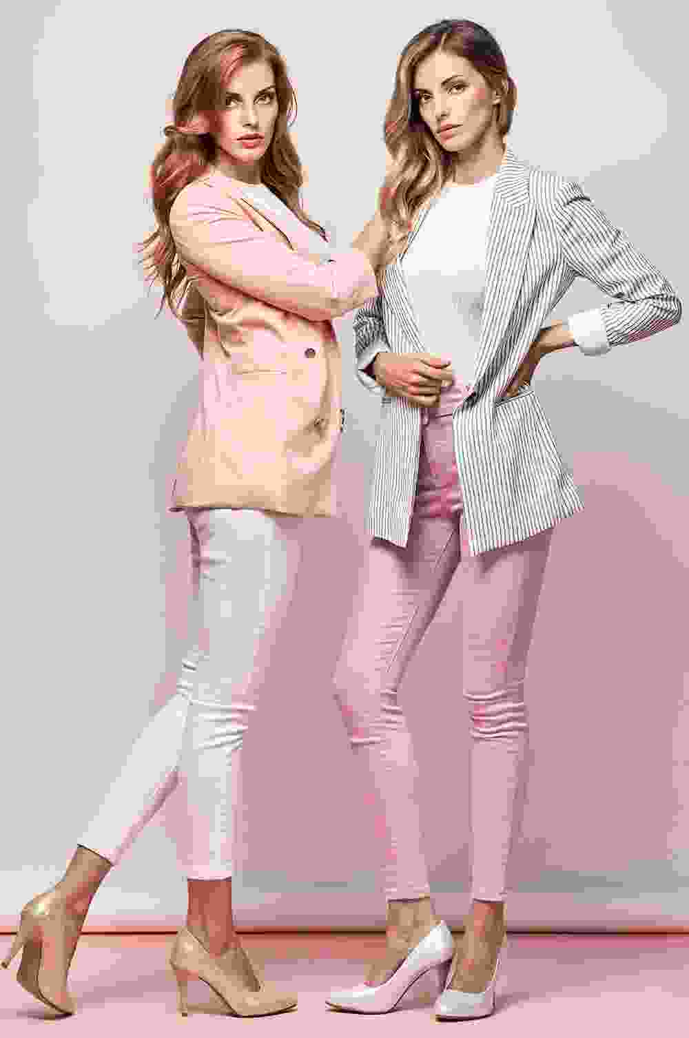 Two stylish women posing confidently in blazers and heels against a pink and white backdrop.