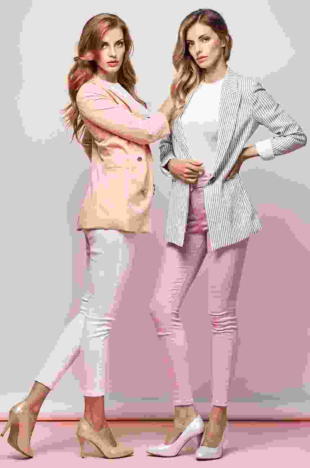 Two stylish women in blazers and heels pose confidently against a pastel backdrop.