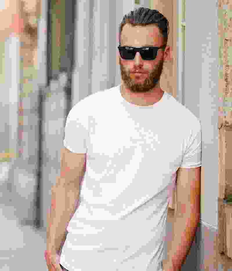 Man with beard and sunglasses wearing a white t-shirt leaning against a wall.