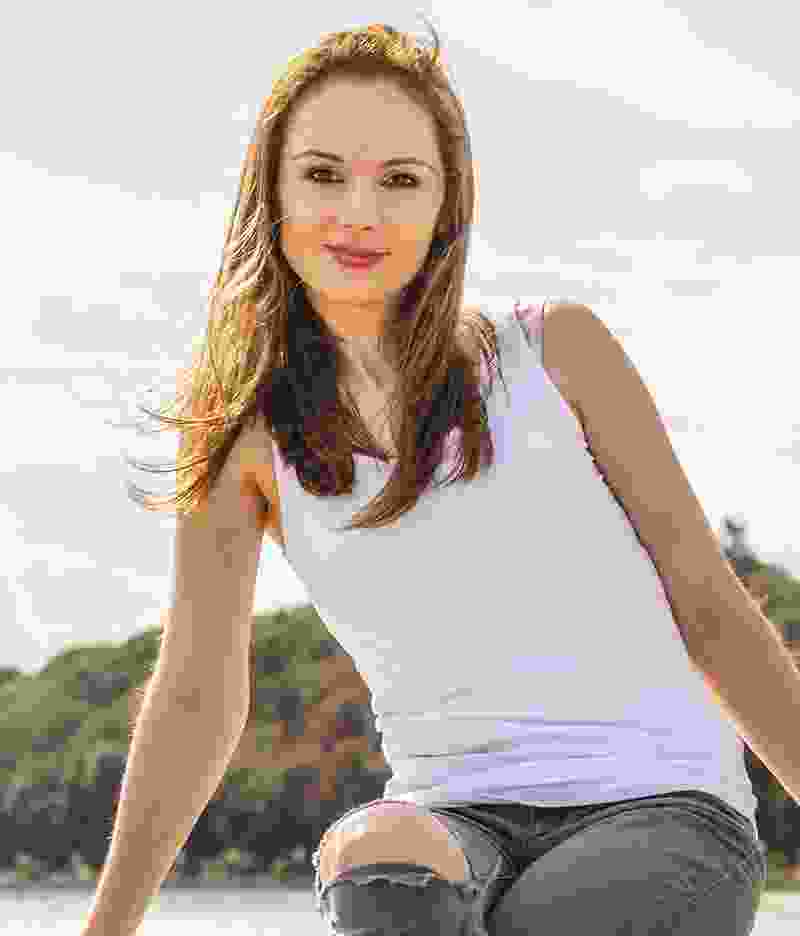 Young woman in white tank top and ripped jeans smiling outdoors.