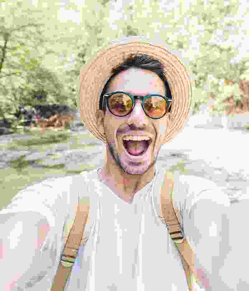A happy man wearing sunglasses and a straw hat takes a selfie outdoors.